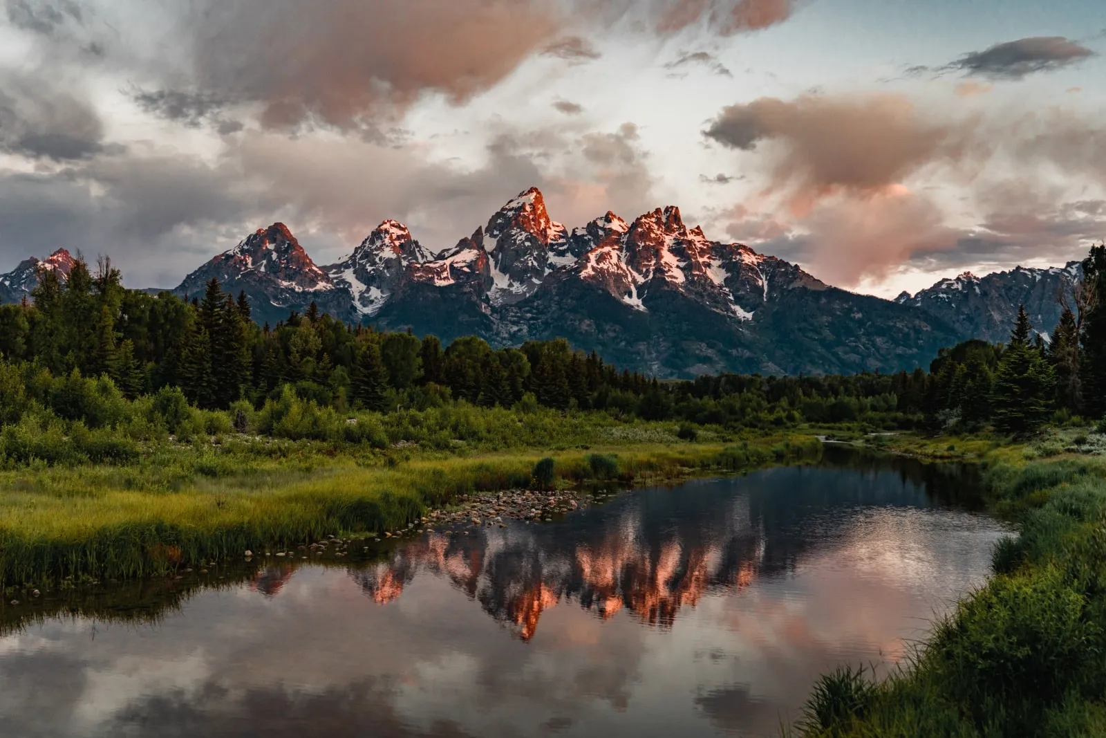A serene picture from the base of the Switchback Loop along a lake with a backdrop of the Grand Teton mountain peaks.